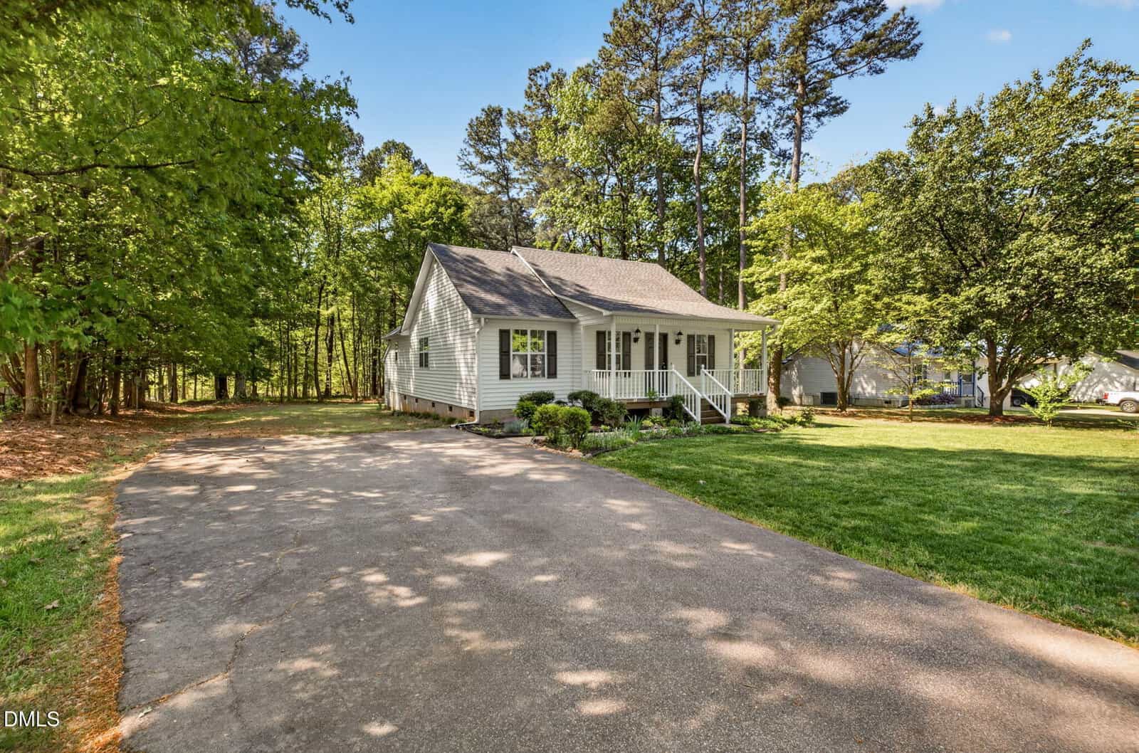 A small white house with a front porch sits beside a long driveway, surrounded by green grass, trees, and a neighborhood under a blue sky.
