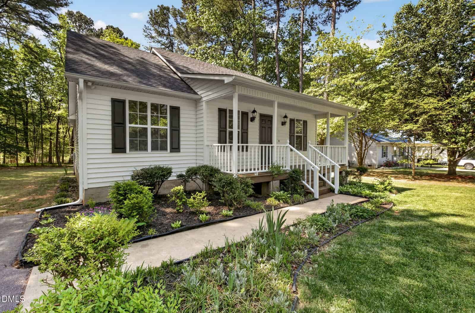 A white single-story house with a covered front porch, dark shutters, and two front doors. The yard has neatly trimmed bushes, flowerbeds, a walkway, and is surrounded by tall trees and green grass.