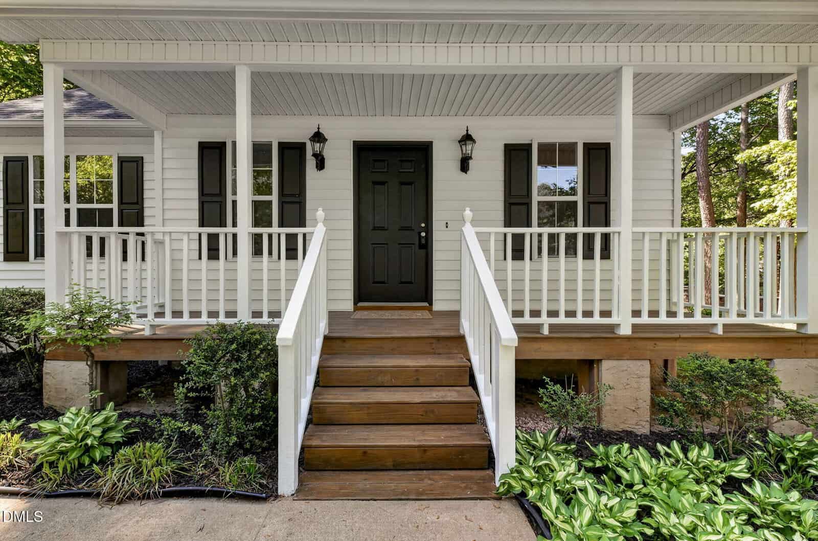 A white house with a covered front porch, black door, black windows, wooden steps, white railing, and landscaped bushes and plants along the walkway.