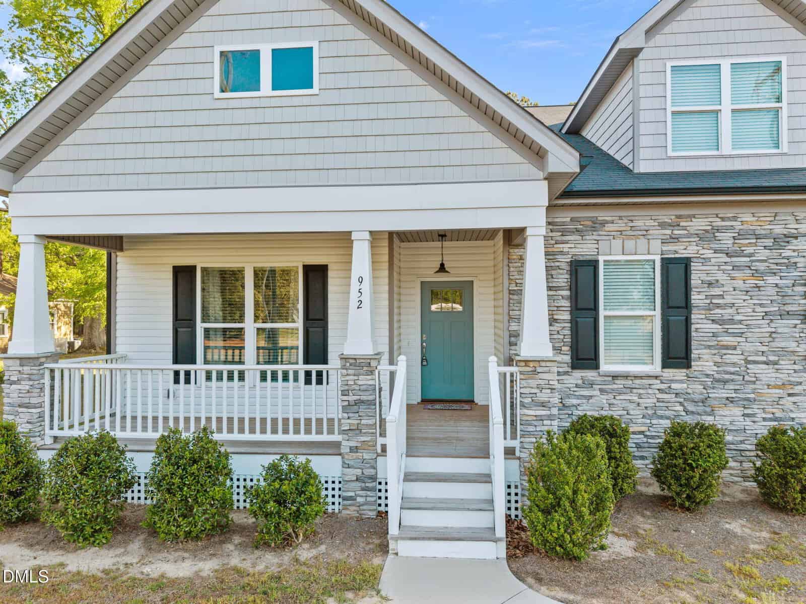 A light gray house with stone accents, white trim, black shutters, a teal front door, and a small covered porch with white railings. Steps lead up to the porch, and trimmed bushes line the front.