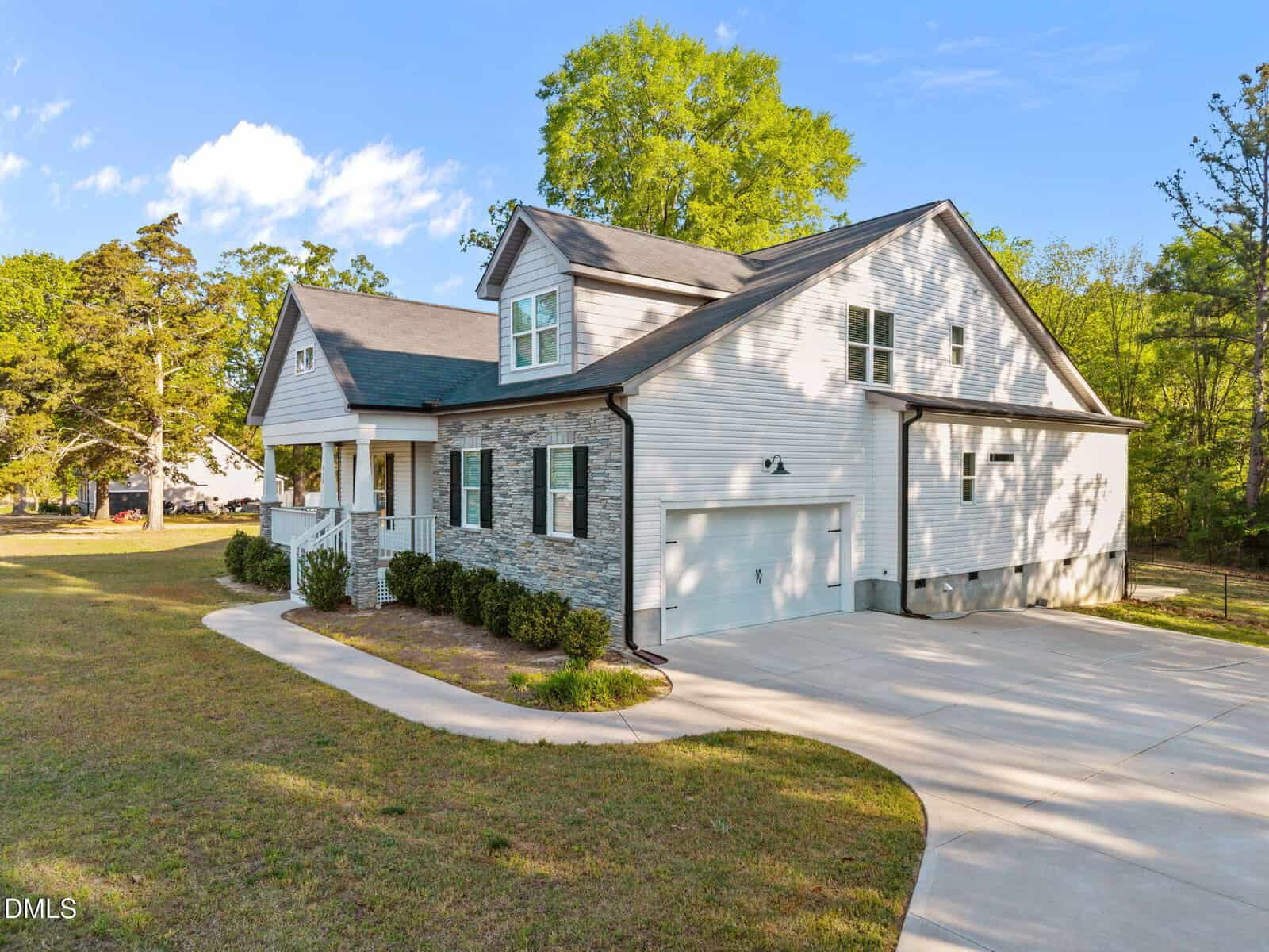 A modern two-story house with light gray siding, stone accents, an attached two-car garage, covered front porch, and a curved driveway, surrounded by grass, trees, and blue sky.
