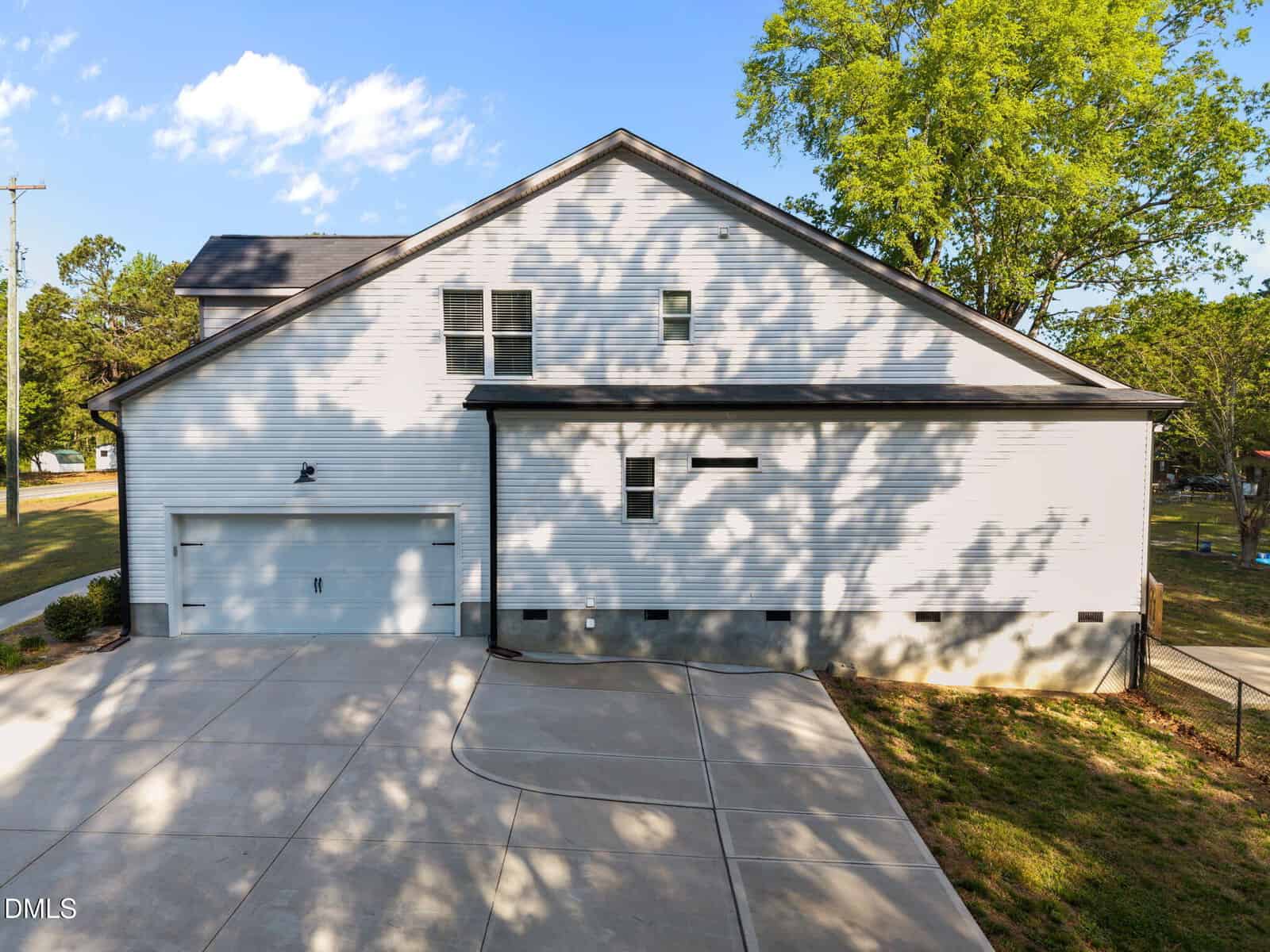A white two-story house with a double garage, large driveway, and shaded by nearby trees under a blue sky with scattered clouds.
