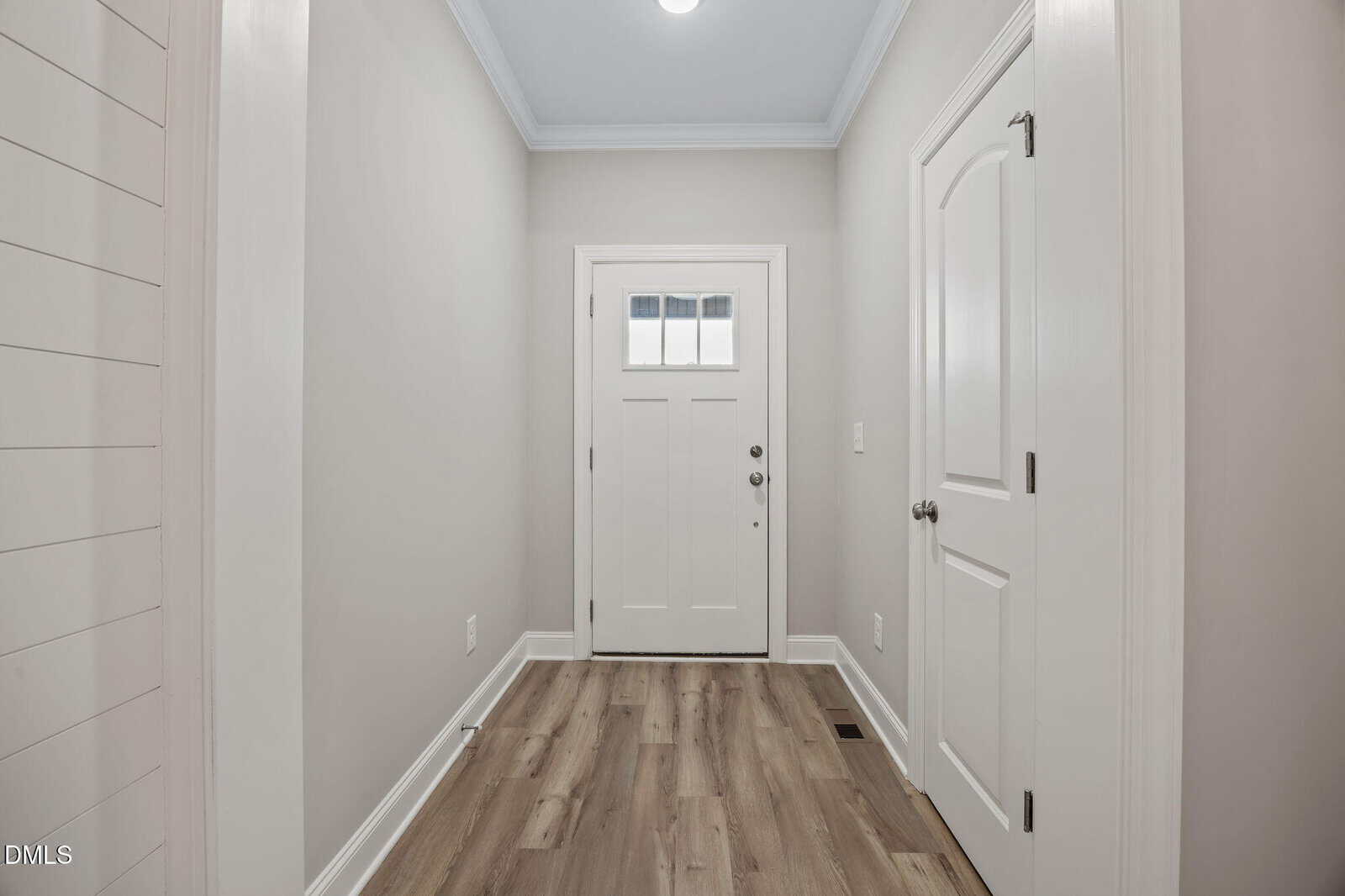 A bright, clean entryway with light wood flooring, a white front door with a small window, and white walls. Two closed white doors are on either side of the hallway.