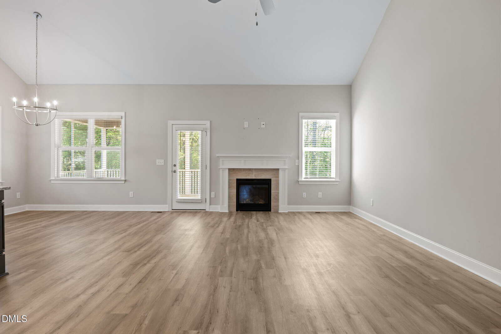 Spacious living room with light wood flooring, a white fireplace centered on the far wall, three windows, a door to a patio, and a modern chandelier to the left. Walls are light gray and ceiling is vaulted.