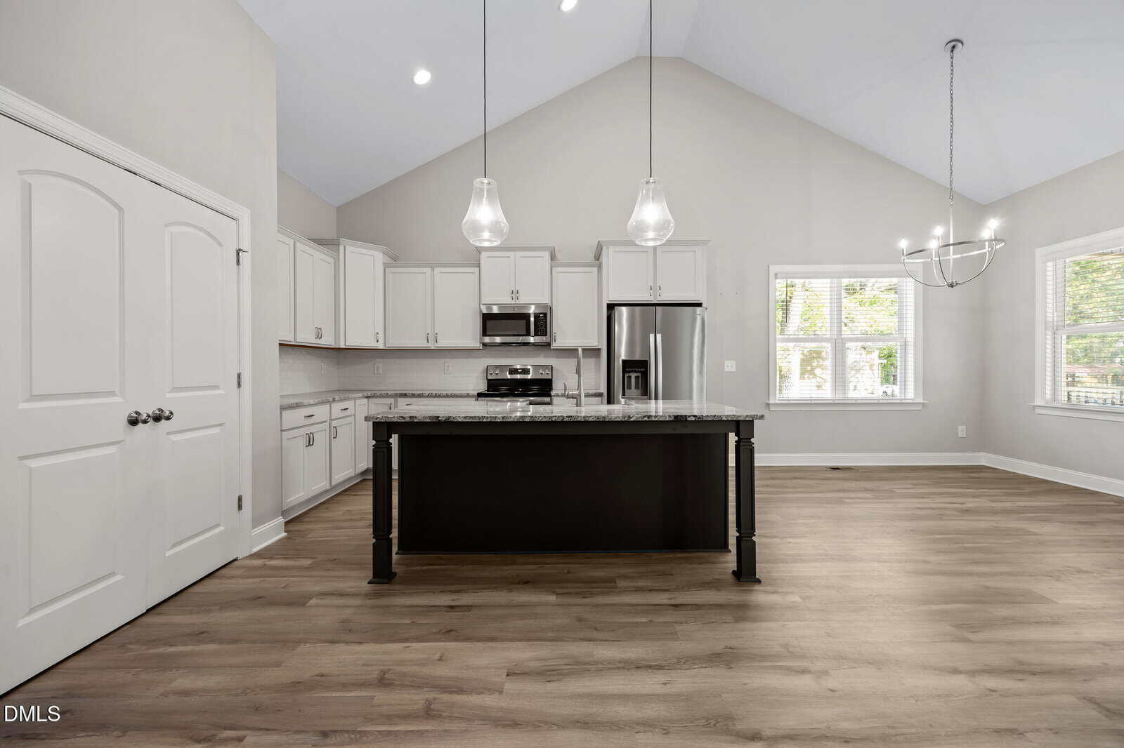 Modern kitchen with white cabinets, stainless steel appliances, a large island with dark base, pendant lights, and light wood flooring. Double doors and windows let in natural light, creating a bright, open space.