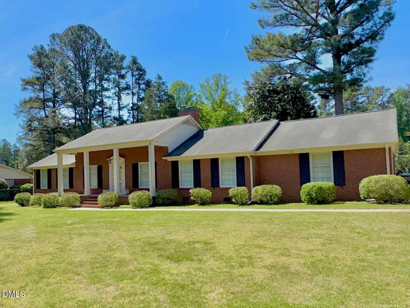 Single-story brick house with white columns, dark shutters, and a large front lawn. Bushes line the front, and tall trees stand in the background under a clear blue sky.