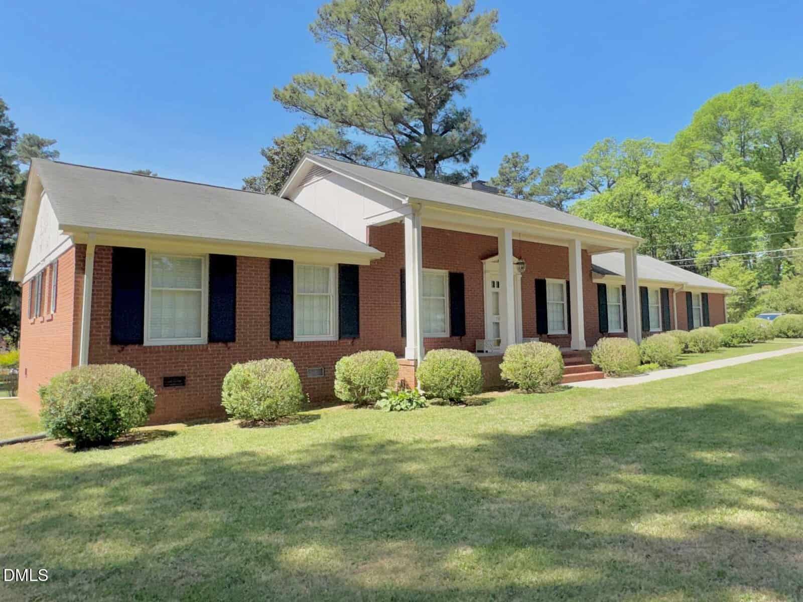 A one-story red brick house with white columns, black shutters, and a covered front porch, surrounded by neatly trimmed bushes and a green lawn, with tall trees in the background under a clear blue sky.