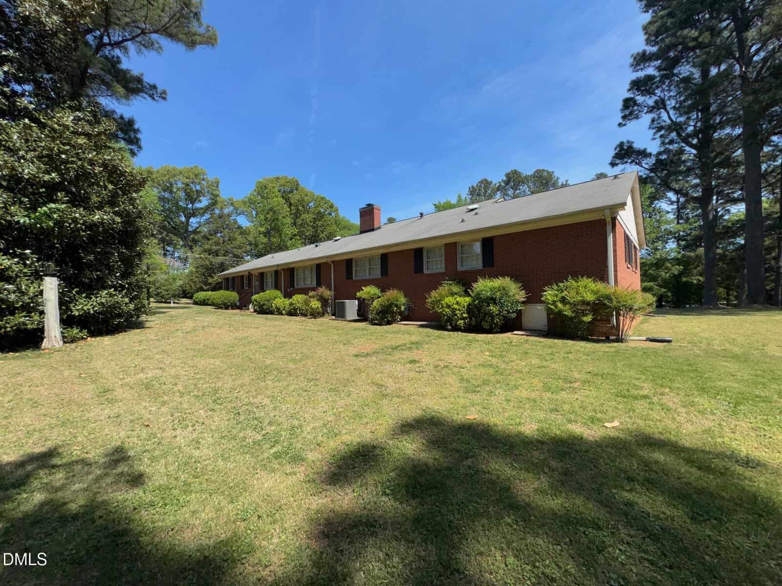 A long, single-story brick house with white trim sits on a large, grassy lawn surrounded by tall trees and bushes, under a clear blue sky.