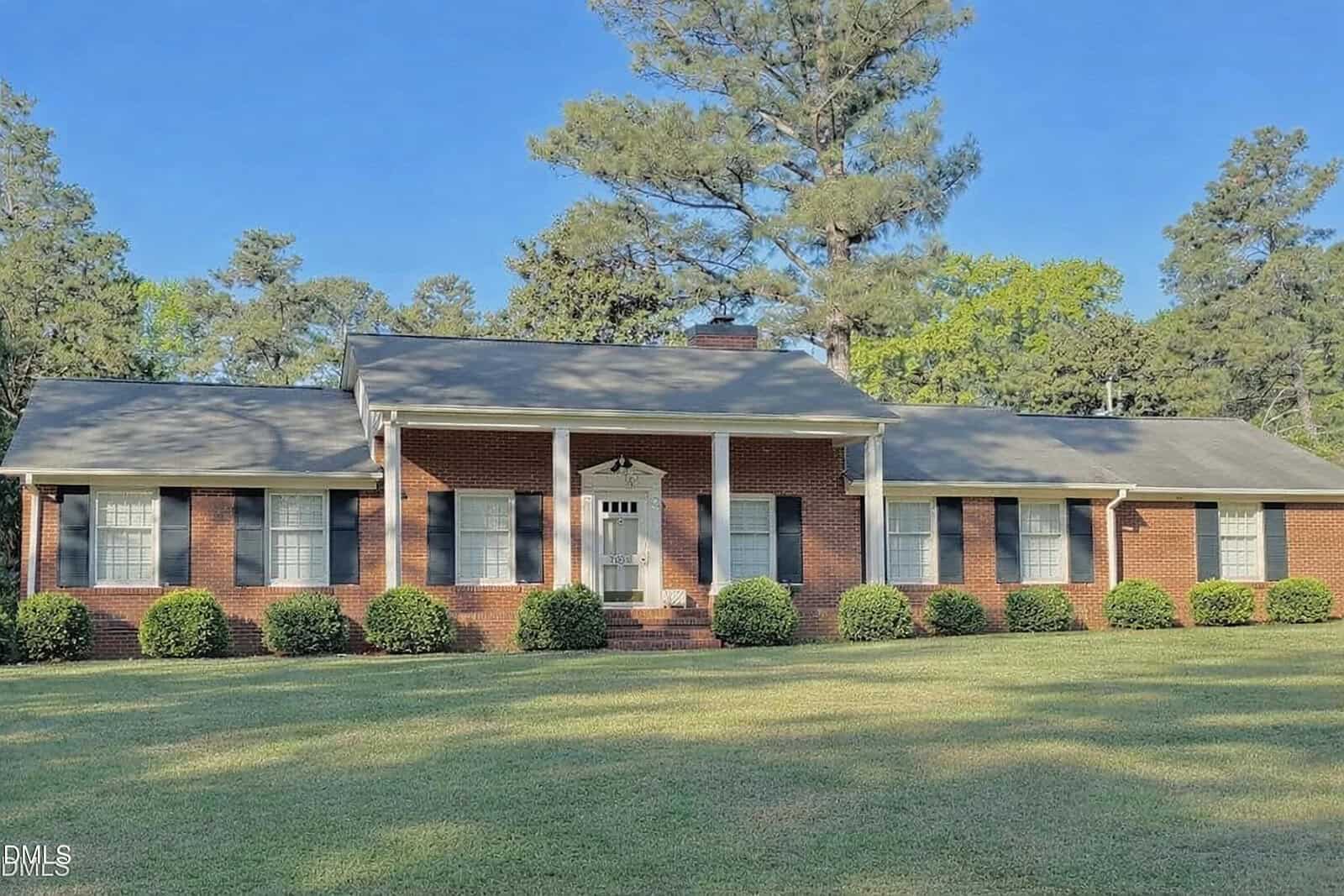 A single-story brick house with white columns, a gabled roof, and symmetrical windows sits on a large grassy lawn at 1604 Crescent Drive, Henderson, surrounded by neatly trimmed bushes and tall pine trees in the background.