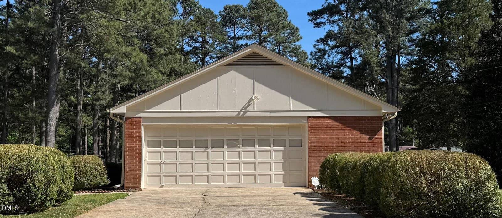 A brick and white two-car garage with a closed door, surrounded by neatly trimmed bushes and tall trees under a clear blue sky.