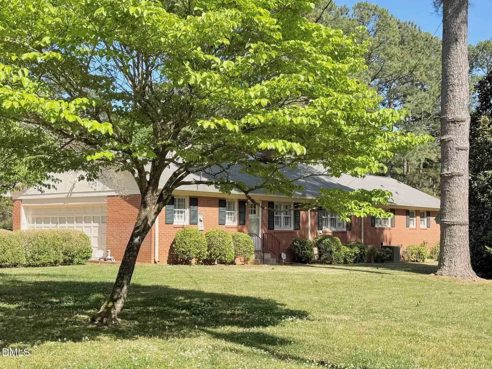 A single-story brick house with white trim and a front porch, surrounded by green bushes and shaded by large leafy trees on a sunny day. The yard is spacious with well-kept grass.