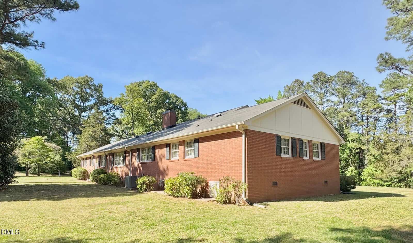 A single-story red brick house with white trim, multiple windows, and a chimney, surrounded by grass, trees, and shrubs, under a clear blue sky.