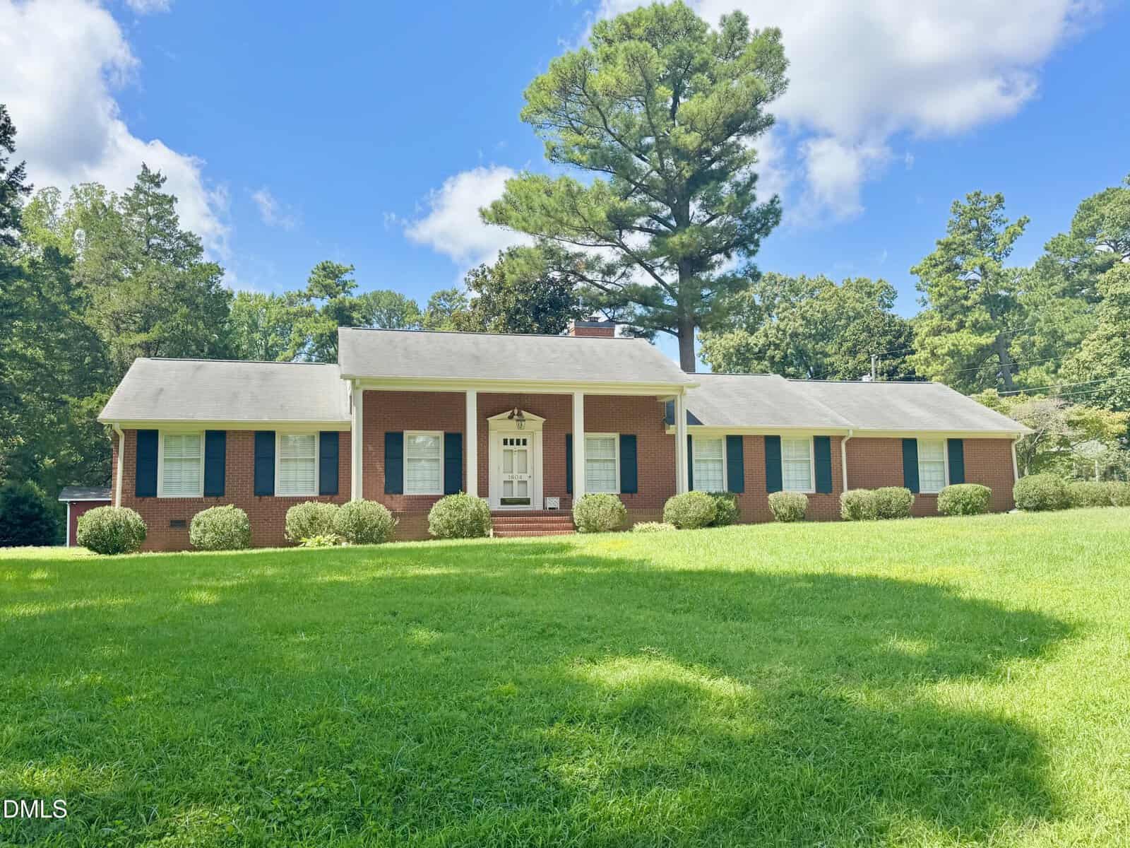 A single-story red brick house with white trim, dark shutters, and a covered front porch sits on a large green lawn surrounded by trees under a partly cloudy sky.