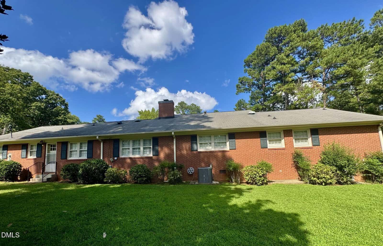 A single-story red brick house with a gray roof, white trim, and black shutters is shown on a sunny day. There are bushes along the front and tall green trees in the background. The sky is blue with scattered clouds.