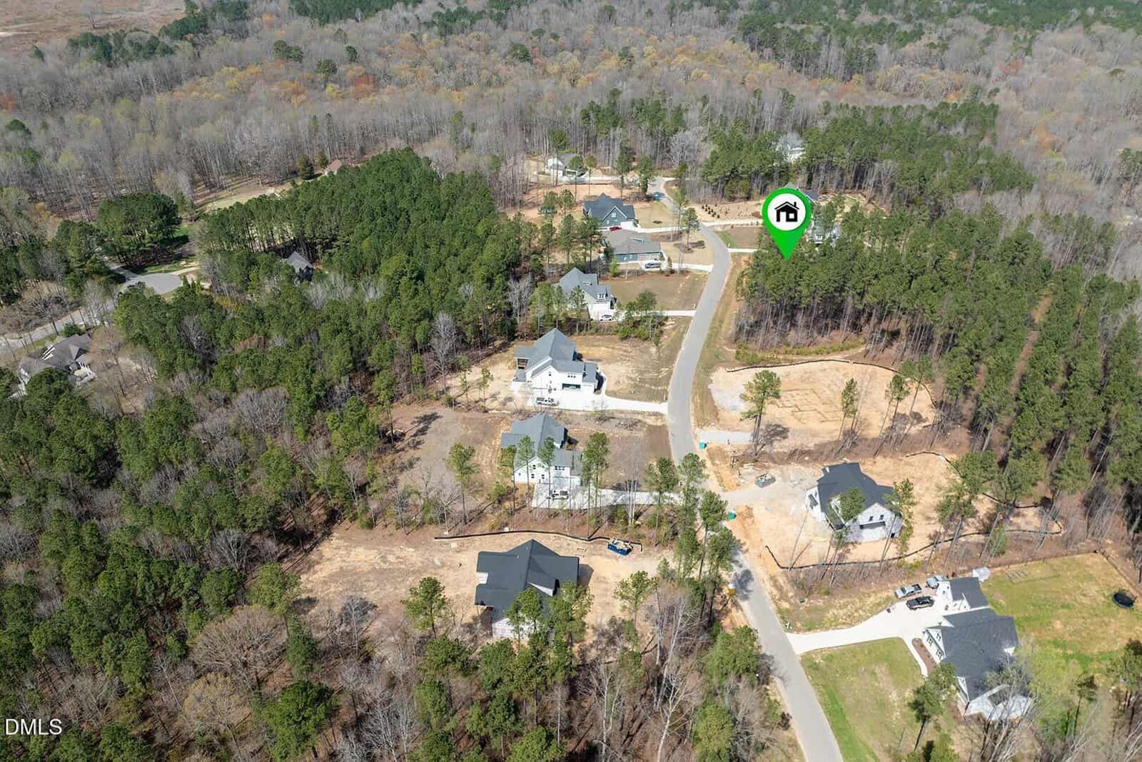 Aerial view of a suburban neighborhood surrounded by trees, with several houses, clearings, and roads. A green location marker with a house icon highlights one property in the upper right area of the image.