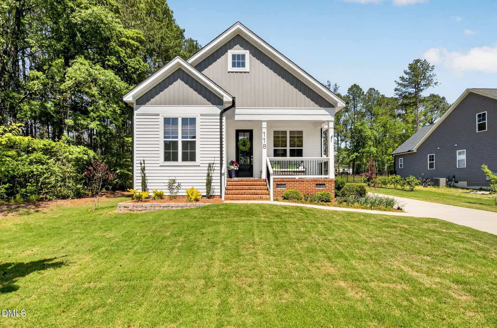 Single-story gray house with white trim, front porch, brick steps, and a small garden. Nestled at 118 Wichita Way in Louisburg, the house is surrounded by a green lawn and trees, with a driveway on the right leading to the backyard.