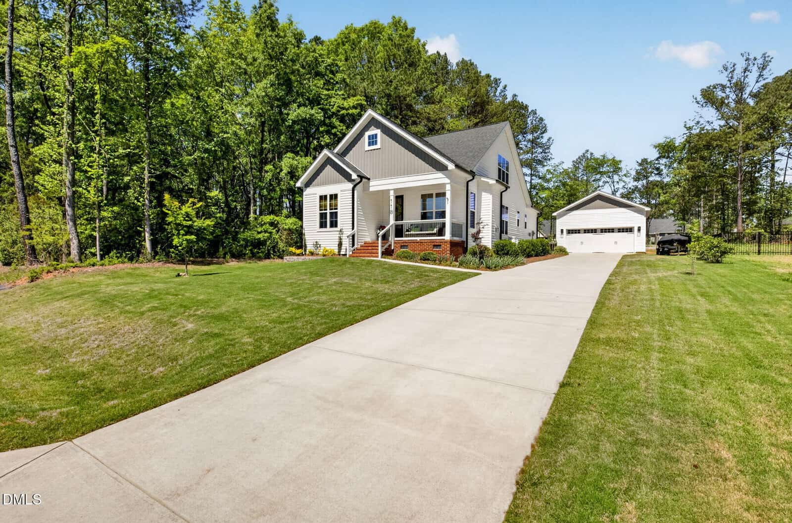 A white house with gray roof sits near trees, with a long concrete driveway leading to a detached garage. The lawn is mowed, and the sky is clear. A vehicle is parked near the garage.