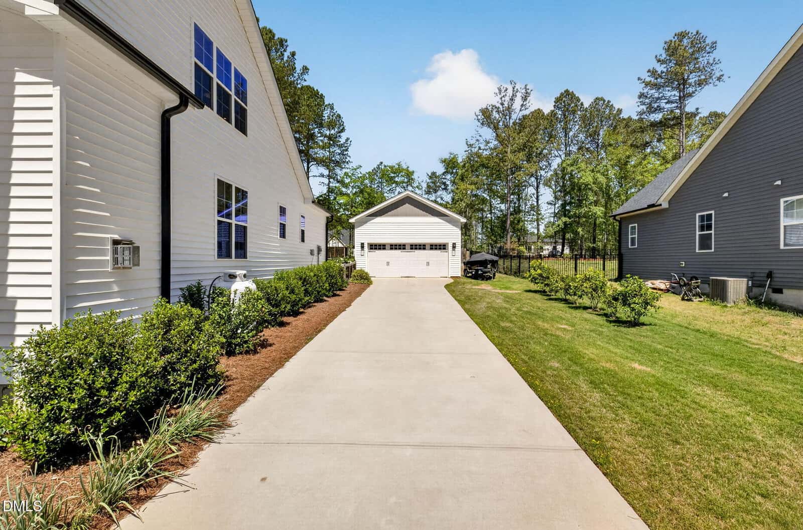 A concrete driveway leads from a white house to a detached two-car garage. Green grass and shrubs are on both sides, with another house visible on the right. Trees and blue sky are in the background.