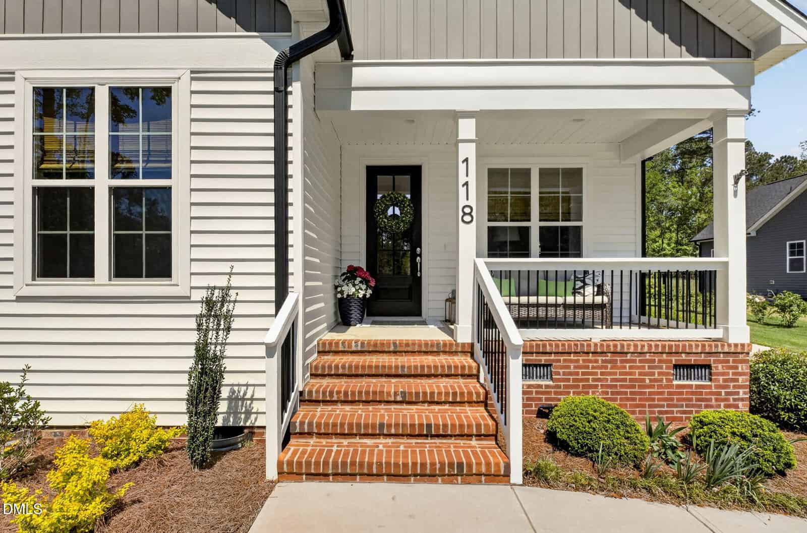 A front porch with brick steps, a black handrail, a black door with a wreath, white siding, the house number 118, and landscaped shrubs and plants along the sidewalk.