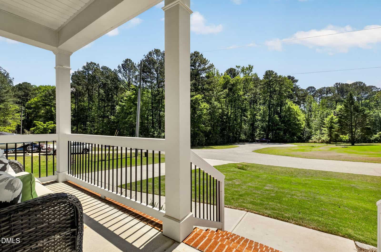View from a covered porch with white railings and brick steps, looking out onto a green lawn, circular driveway, and trees under a clear blue sky. Wicker furniture sits in the left corner of the porch.
