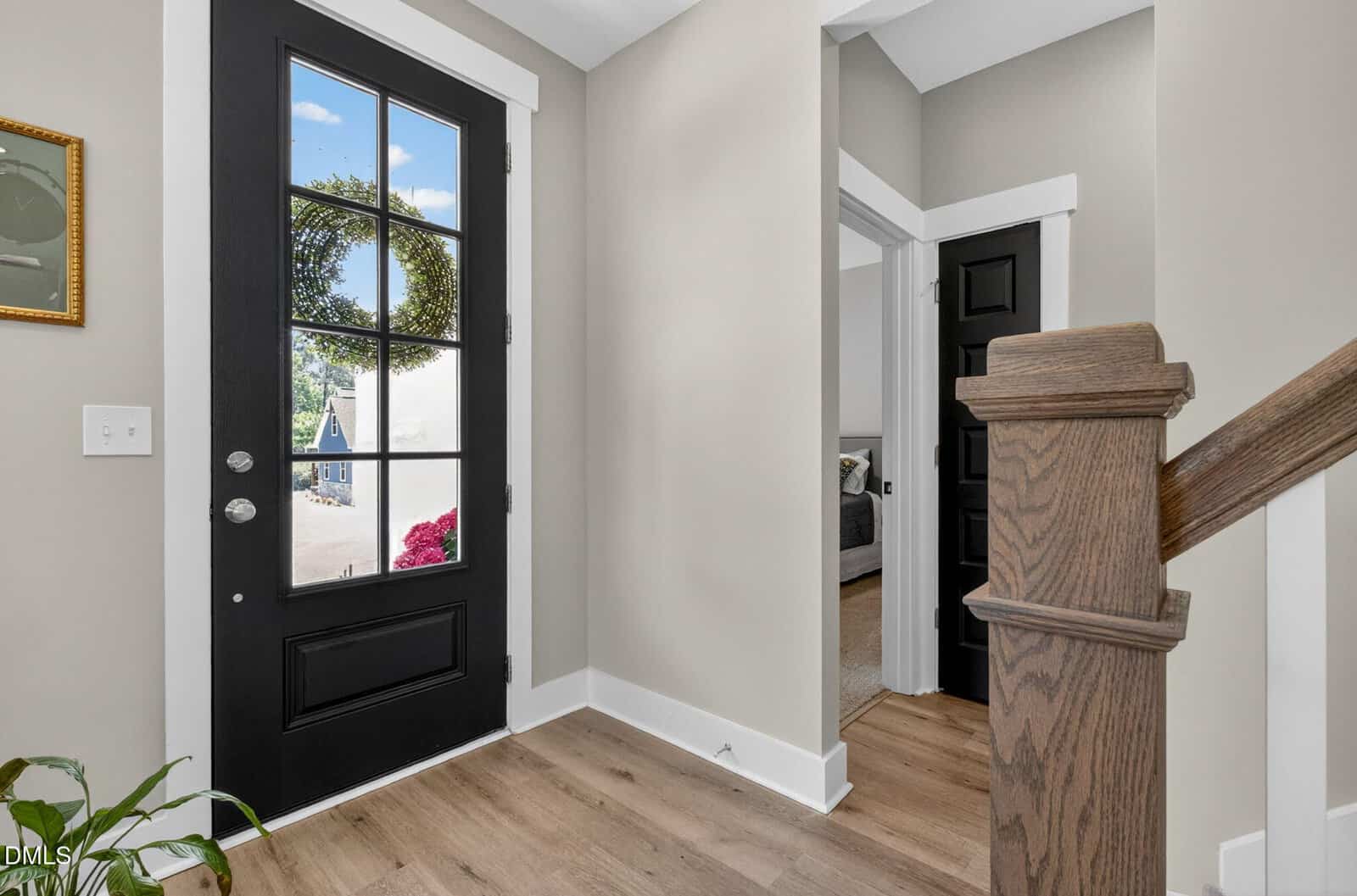 Modern entryway with a black front door, wood floors, white trim, a staircase with a wooden banister, and a glimpse into a bedroom. Sunlight streams through the glass door, showing greenery and houses outside.