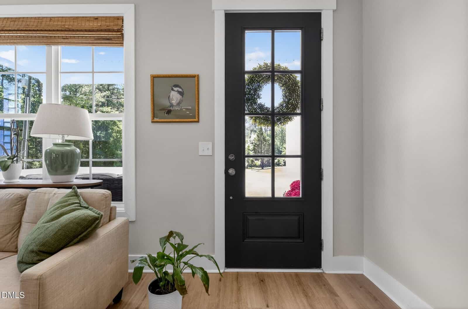 A black front door with a wreath, next to a sofa with green pillows, a lamp, a houseplant, and a wall picture of a bird; sunlight streams through large windows.