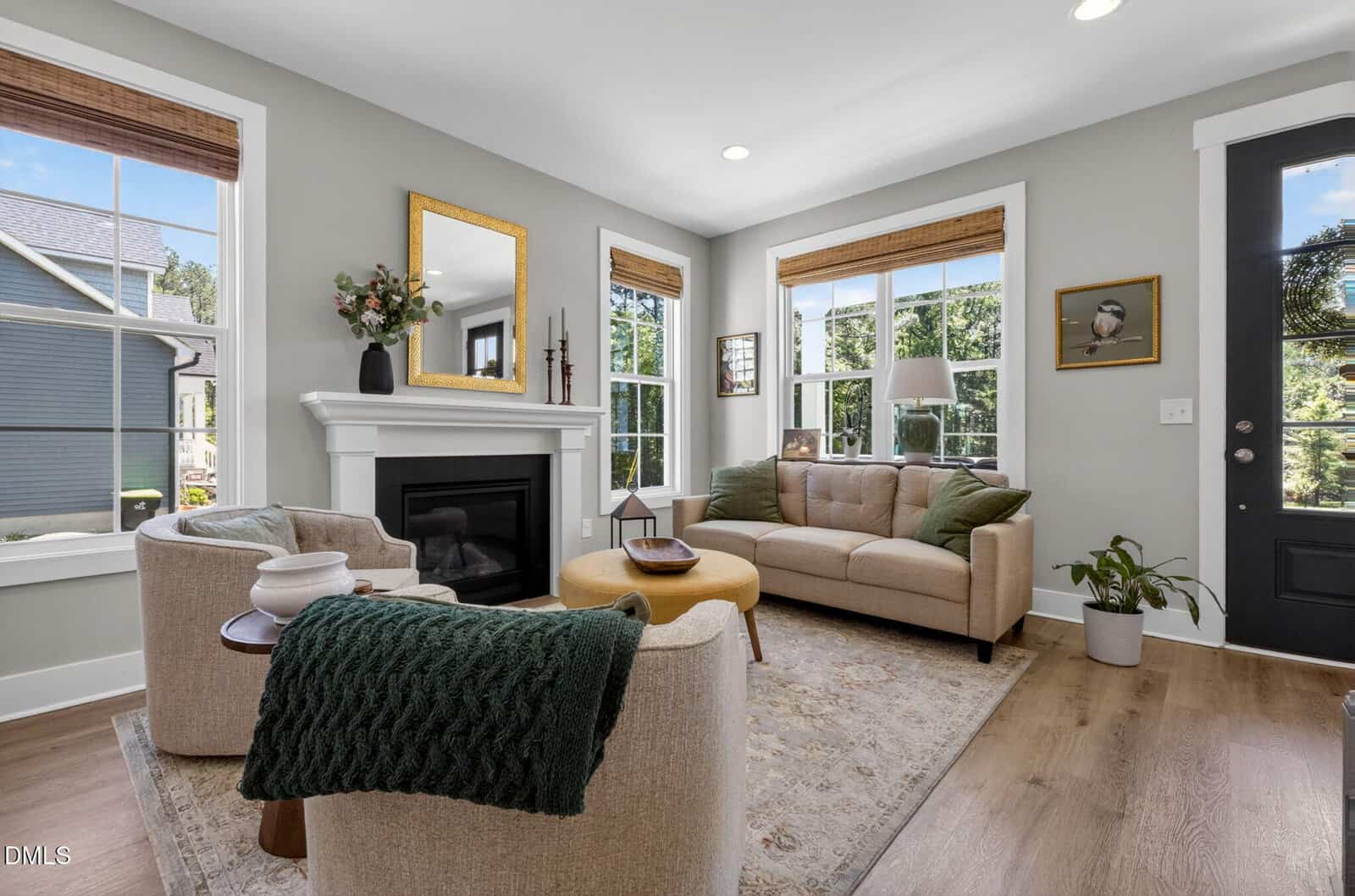 Bright living room with a beige sofa, armchair, fireplace, and windows letting in natural light. Green throw blanket, round ottoman, potted plant, and framed art add cozy, modern touches to the space.