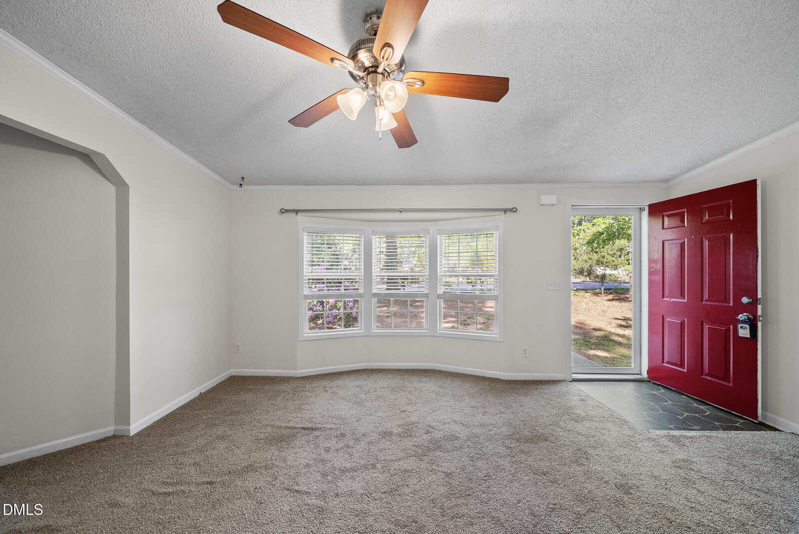 A carpeted living room with a ceiling fan, bay window, and an open red front door next to a glass panel, letting in natural light. White walls and crown molding complete the space.