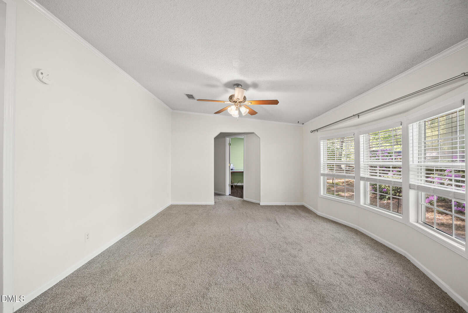 Spacious carpeted room with cream walls, ceiling fan, large bay window with blinds, and arched doorway leading to another room with green walls. Natural light fills the space.