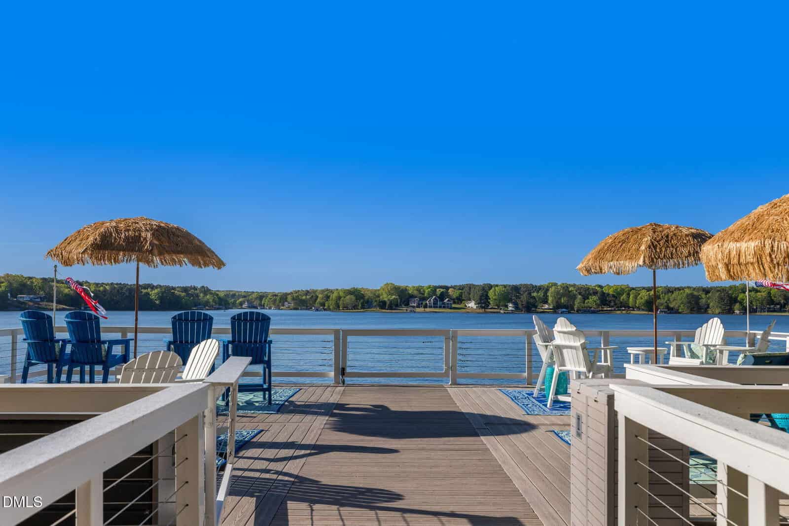 A lakeside deck with blue and white Adirondack chairs, tiki umbrellas, and a clear blue sky overlooking calm water and a tree-lined shore in the distance.