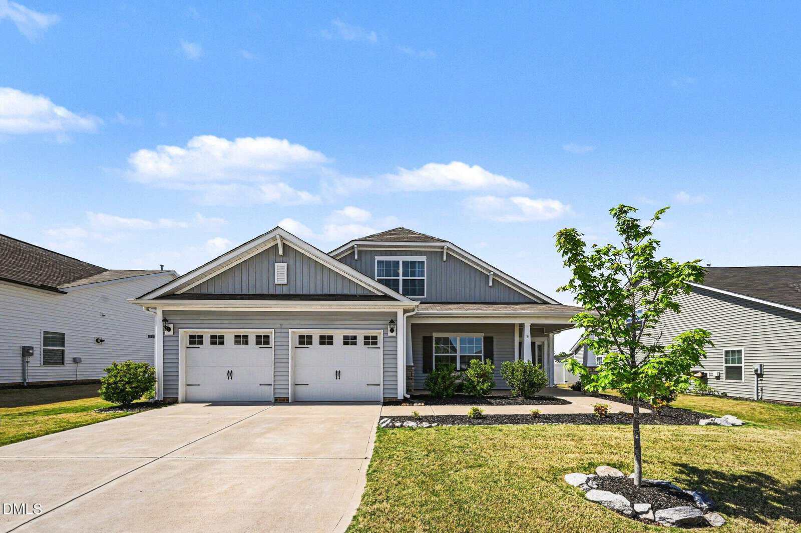 A modern, light gray house with a double garage, neatly landscaped yard, and a small tree in the front. The sky is bright with scattered clouds, and neighboring houses are visible on either side.