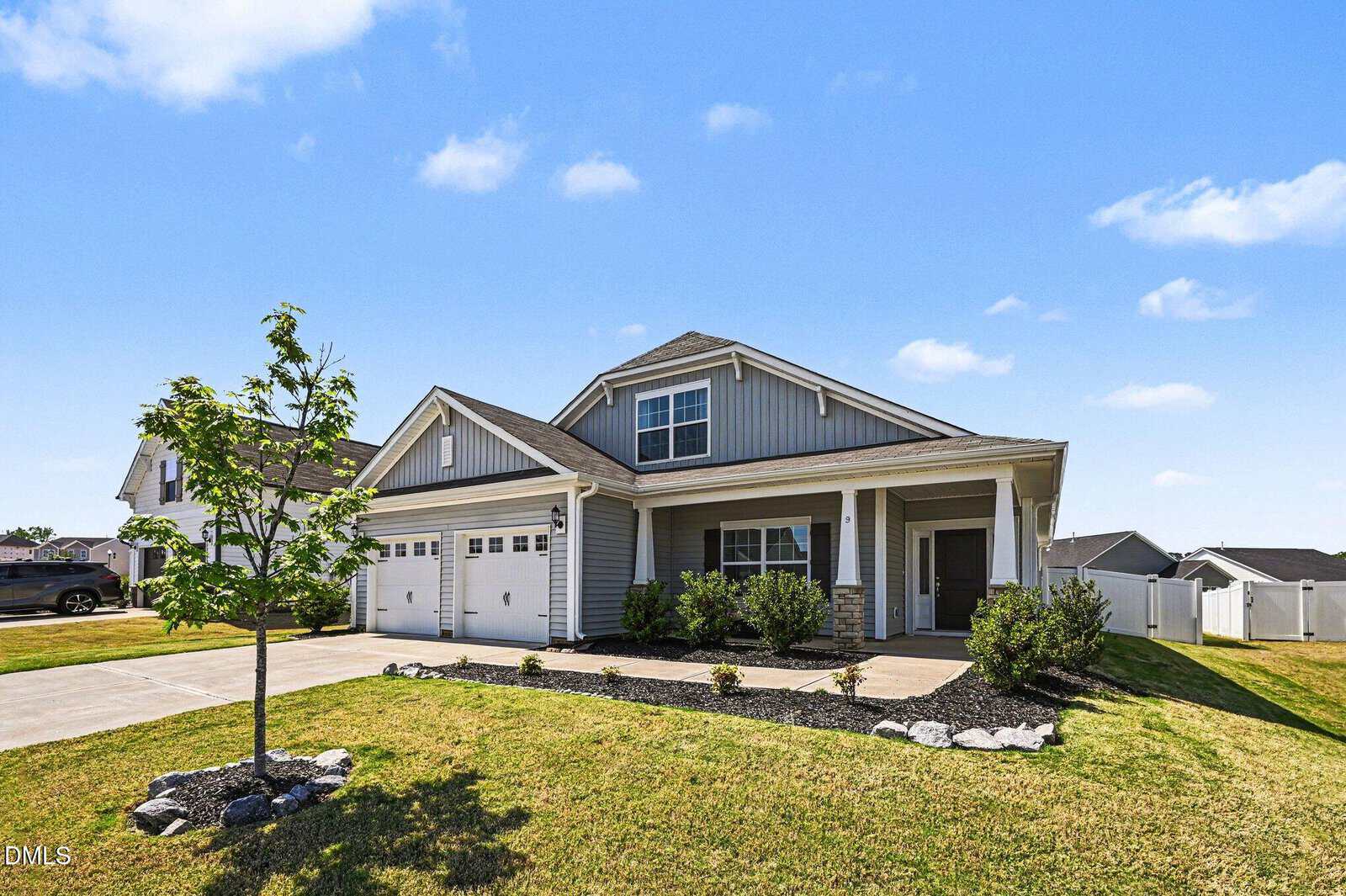 A modern two-story house with gray siding, white trim, a front porch, attached double garage, and landscaped yard with a small tree and shrubs under a sunny blue sky.