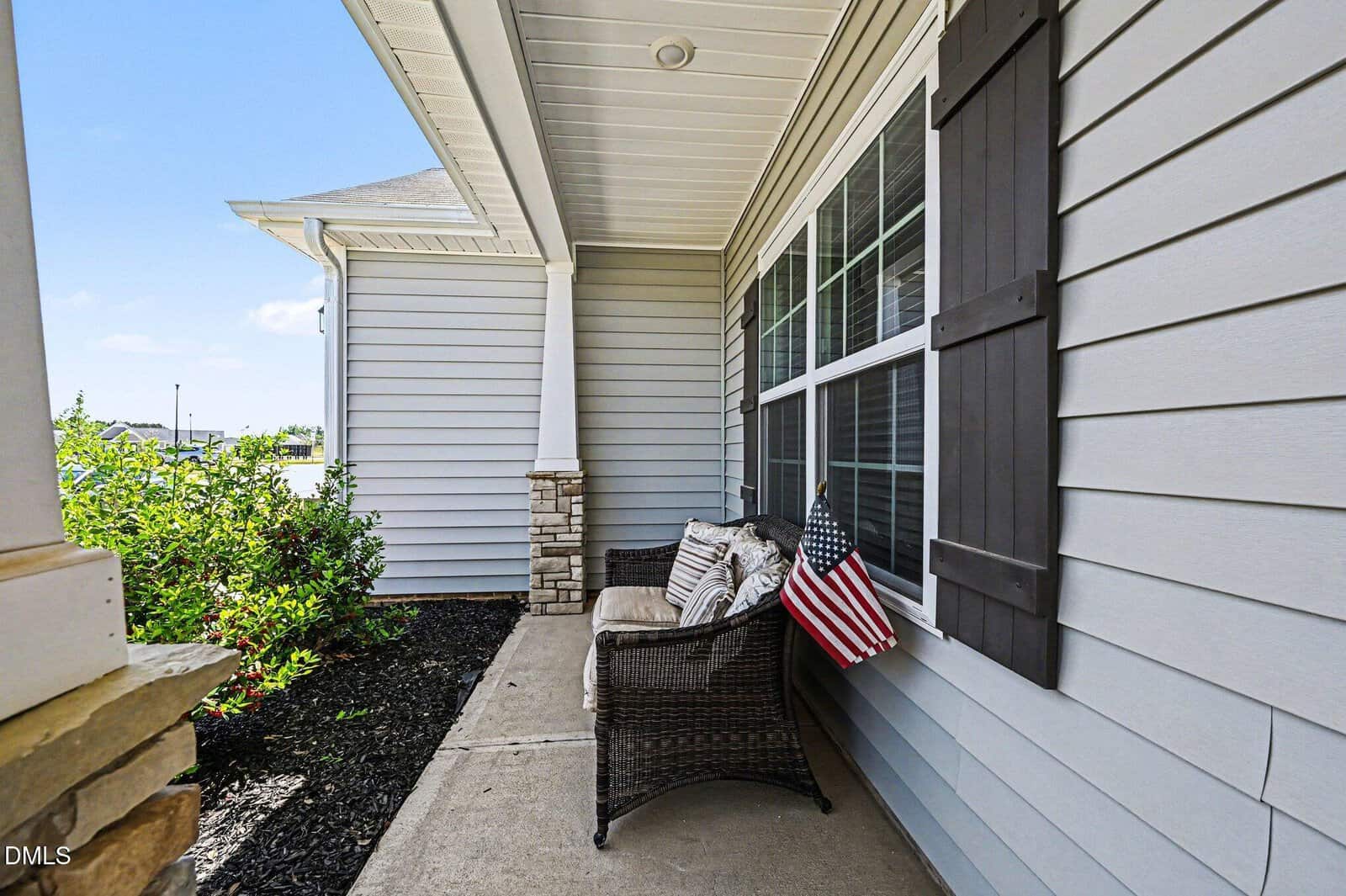 A cozy porch with gray siding and stone accents, featuring a wicker bench with cushions and a small American flag, overlooks landscaped shrubs under a bright blue sky.