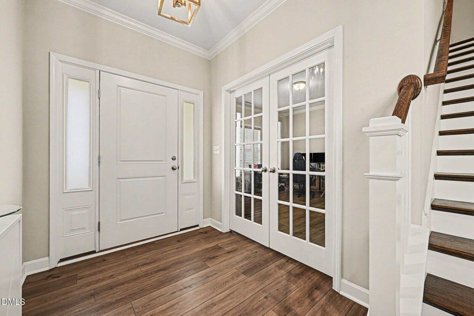 Bright entryway with wood flooring, white front door flanked by narrow windows, glass double doors leading to another room, and staircase with wooden handrail on the right.