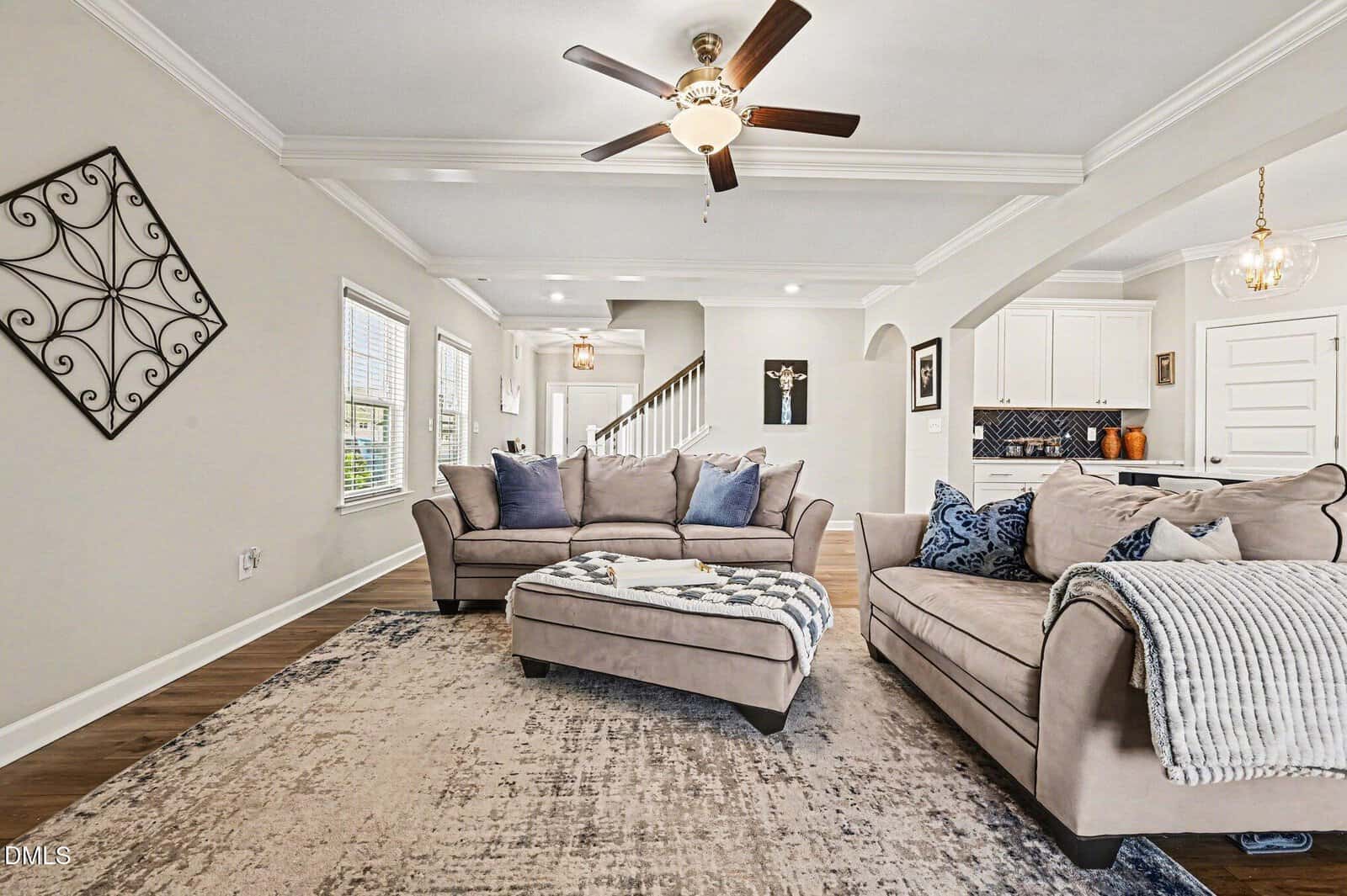 Modern living room with two beige sofas, a matching ottoman, a patterned rug, ceiling fan, and wall art. Bright windows and an open layout lead to a white kitchen and staircase.