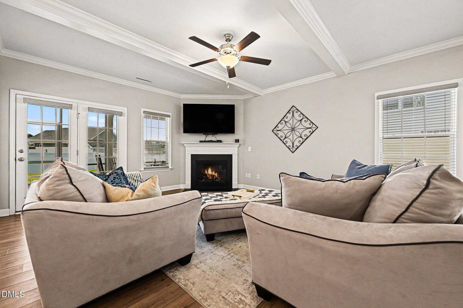 A modern living room with two beige sofas, a patterned ottoman, a fireplace with a TV above it, hardwood floors, large windows, and a ceiling fan with lights. Neutral tones and decorative pillows add comfort.