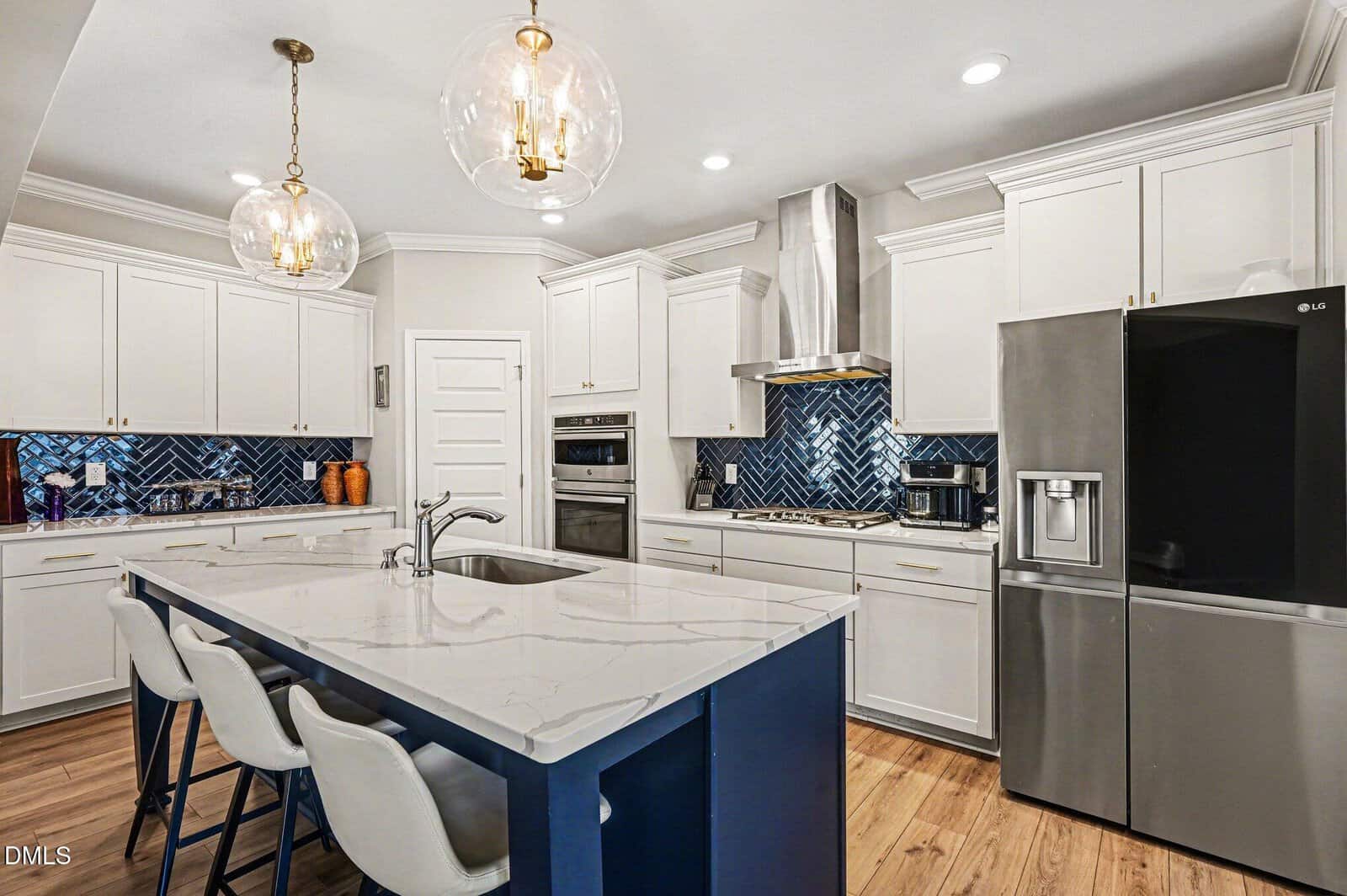 Modern kitchen with white cabinets, blue herringbone backsplash, stainless steel appliances, a marble island with a blue base, pendant lights, and three white bar stools on a wooden floor.