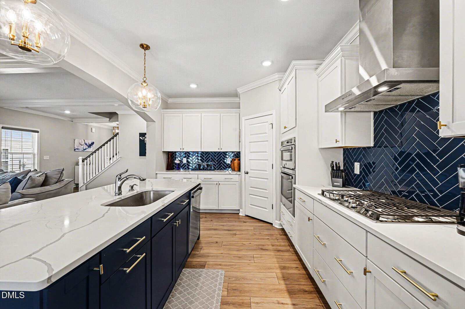 Modern kitchen with white upper cabinets, navy lower cabinets, marble countertops, stainless steel appliances, a blue herringbone backsplash, pendant lights, and wood floors. An open living area and staircase are visible in the background.