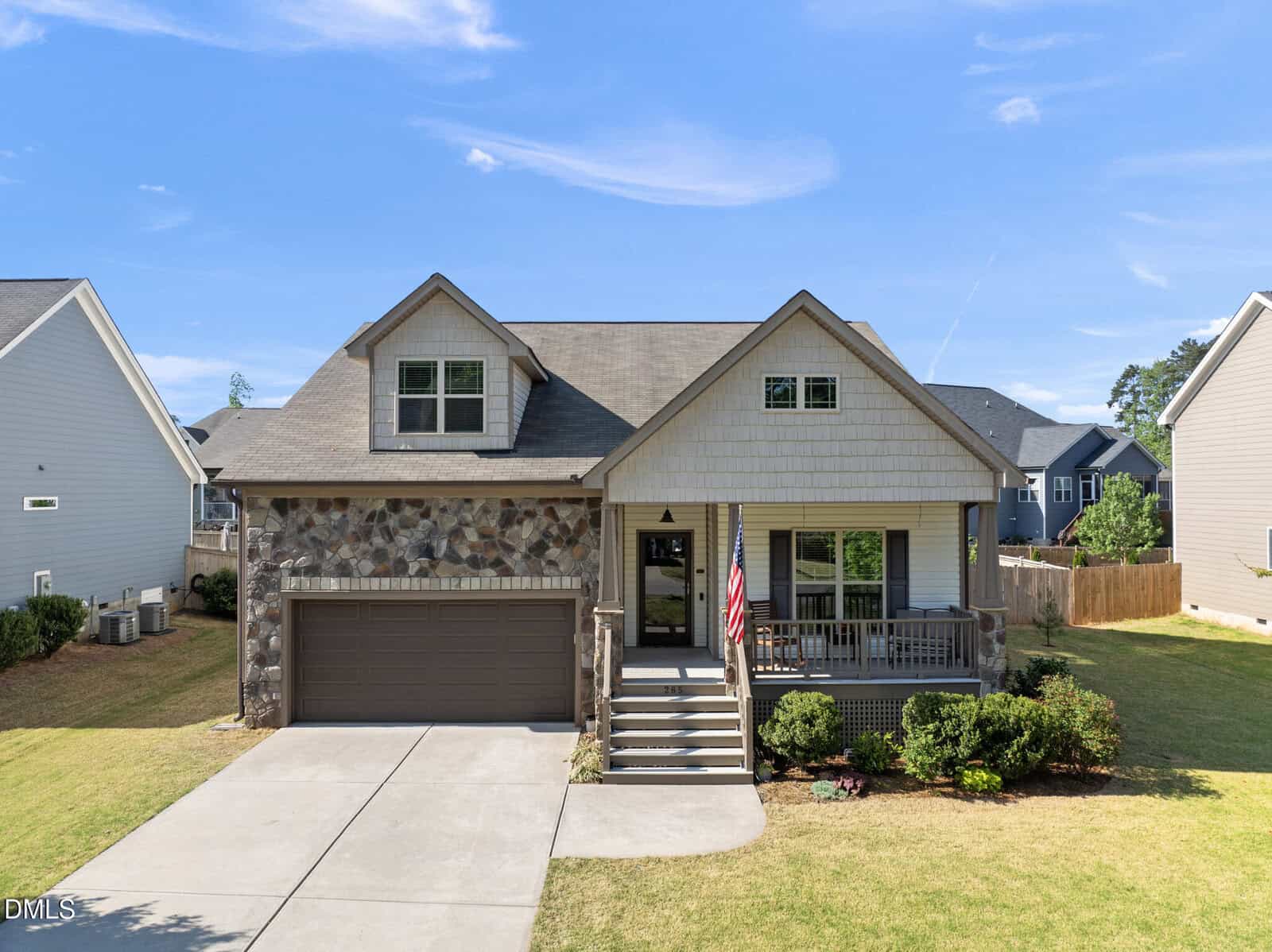 Two-story house with stone and siding exterior, a covered front porch, attached two-car garage, and neatly landscaped yard under a blue sky. An American flag hangs by the porch stairs at 265 Paddy Lane in Youngsville.