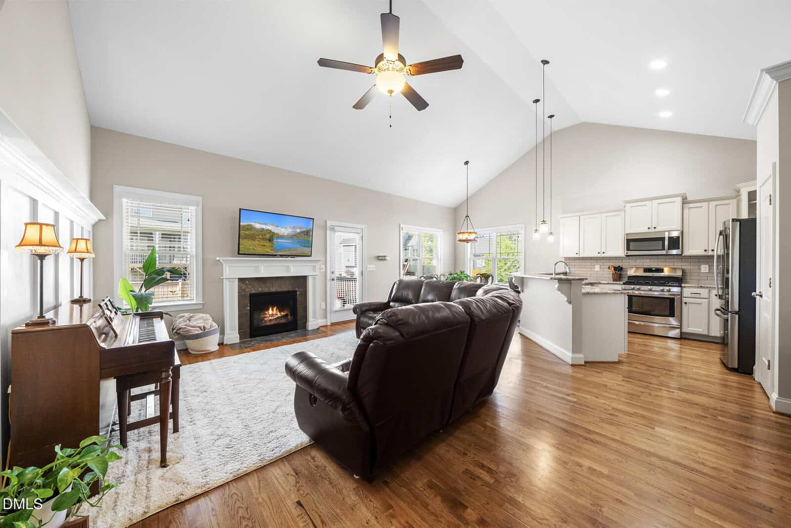 Open-concept living room and kitchen with vaulted ceiling, hardwood floors, brown leather sofas, fireplace, TV, piano, hanging lights, and white cabinetry. Large windows provide natural light.
