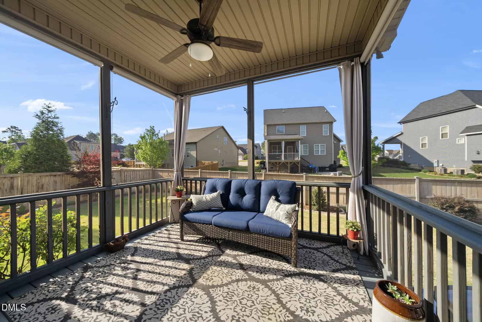 Screened-in porch with a wicker sofa, blue cushions, patterned rug, potted plants, ceiling fan, and a view of a fenced backyard and neighboring houses under a clear blue sky.