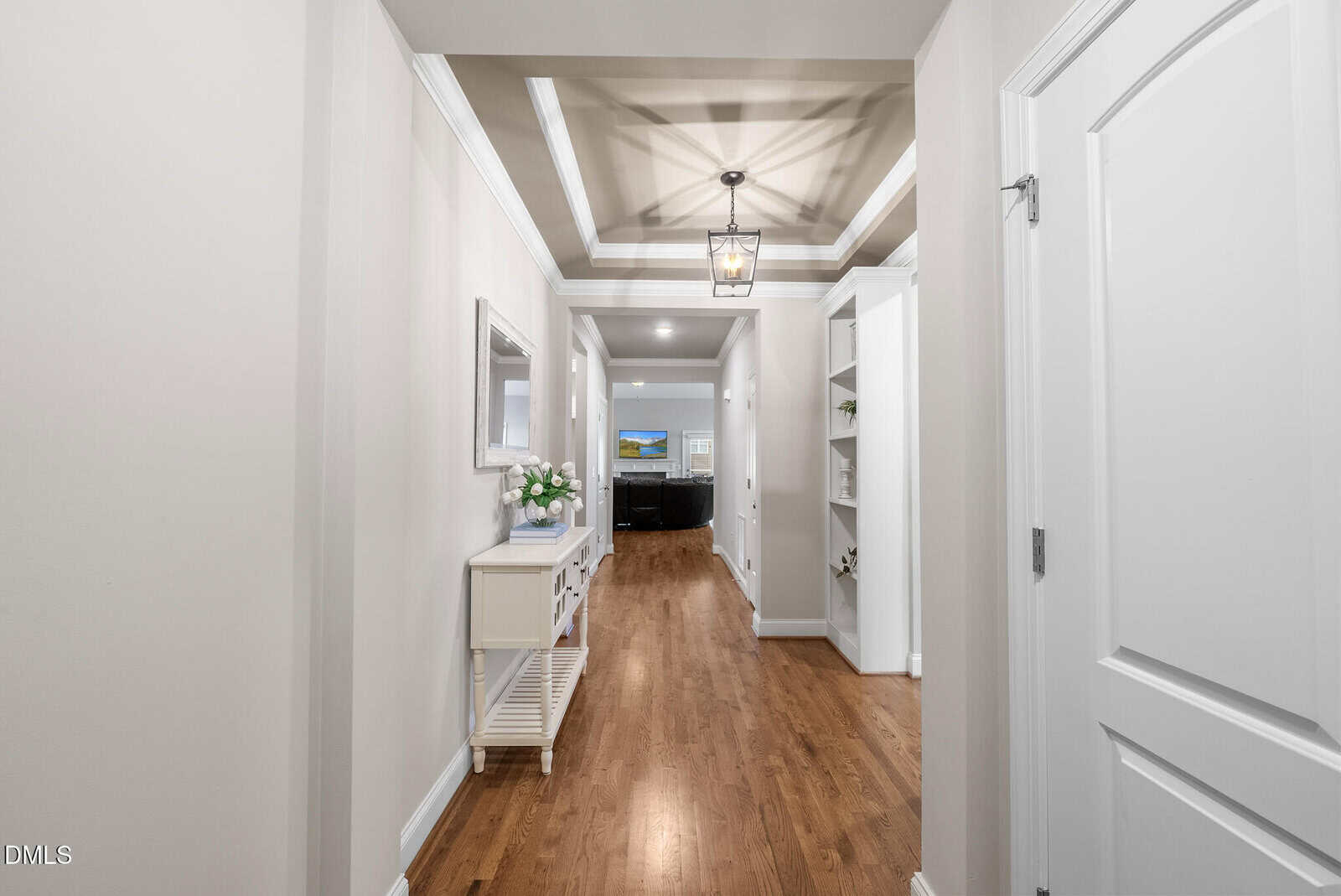 A bright hallway with hardwood floors, white walls, and a tray ceiling leads to a living room. A console table with flowers and a mirror is on the left, and built-in shelves are on the right.