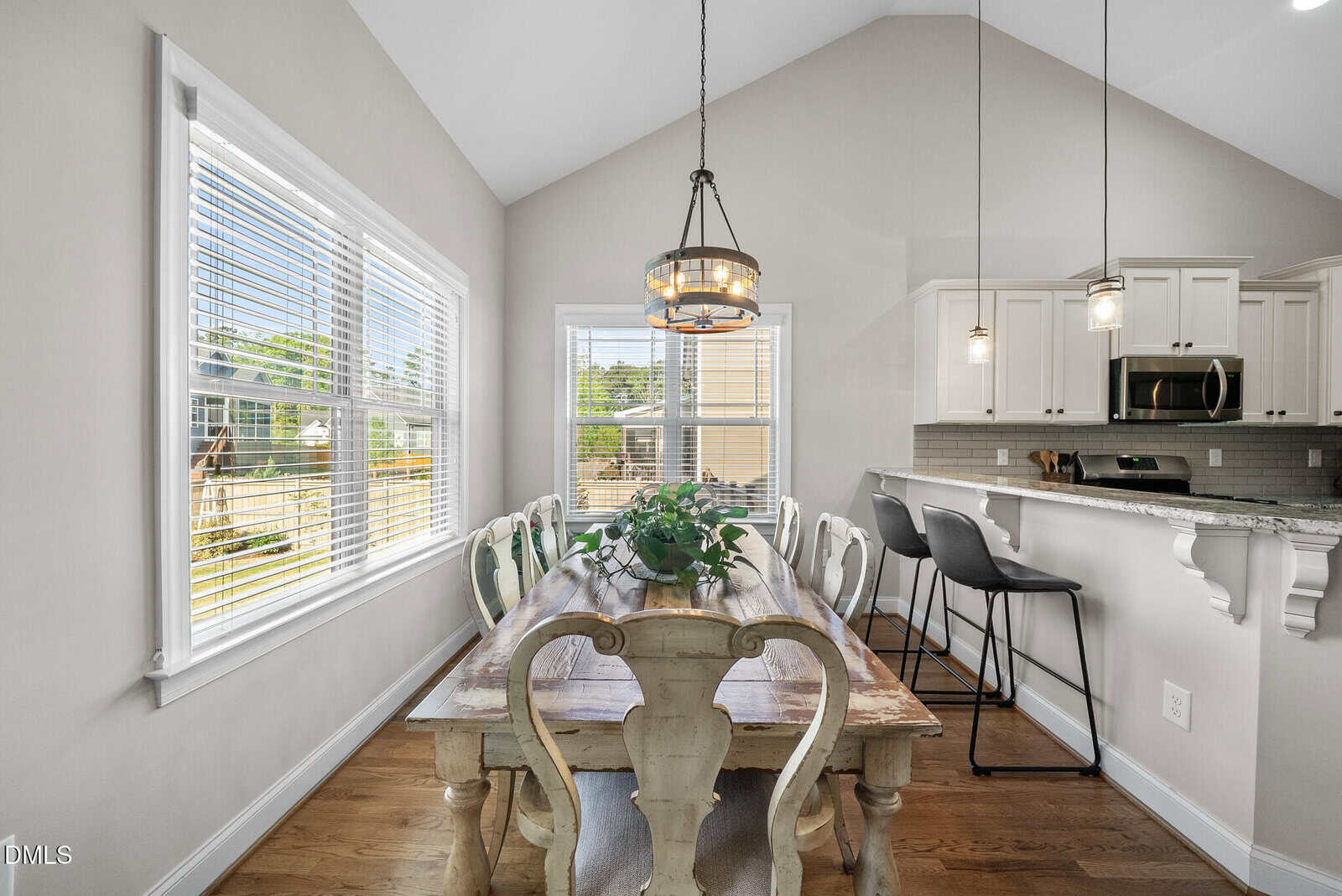 Bright dining area with a rustic wooden table, six chairs, a plant centerpiece, and pendant lights. Kitchen with bar seating and white cabinets is visible, along with large windows letting in natural light.