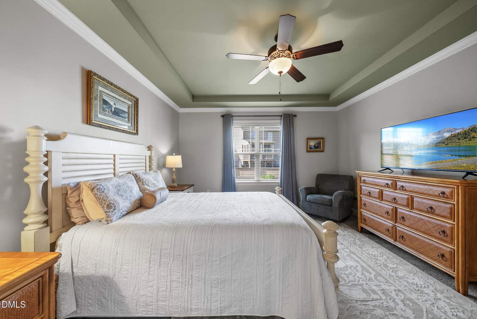 A bright bedroom with a large bed, white bedding, wooden furniture, a ceiling fan, a TV on a dresser, and a window letting in natural light. The room has green tray ceilings and neutral-colored walls.