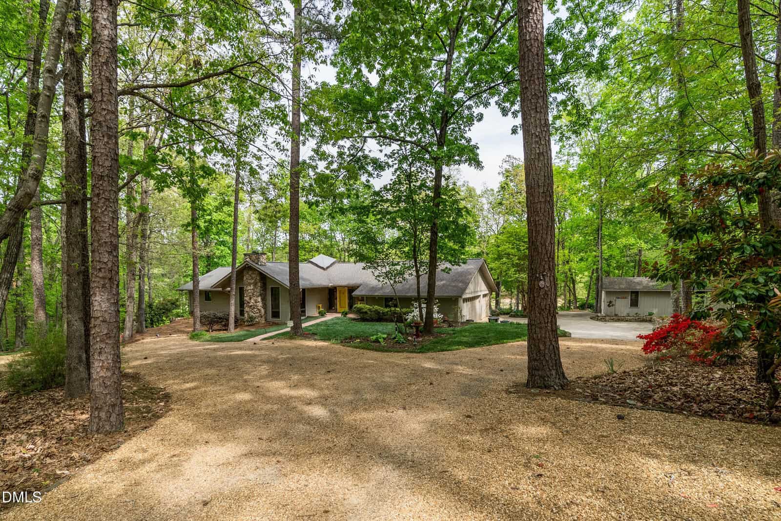 A single-story house with a stone exterior sits among tall trees, with a curved gravel driveway leading to the entrance. The surrounding area is lush and green, with a detached small building in the background.