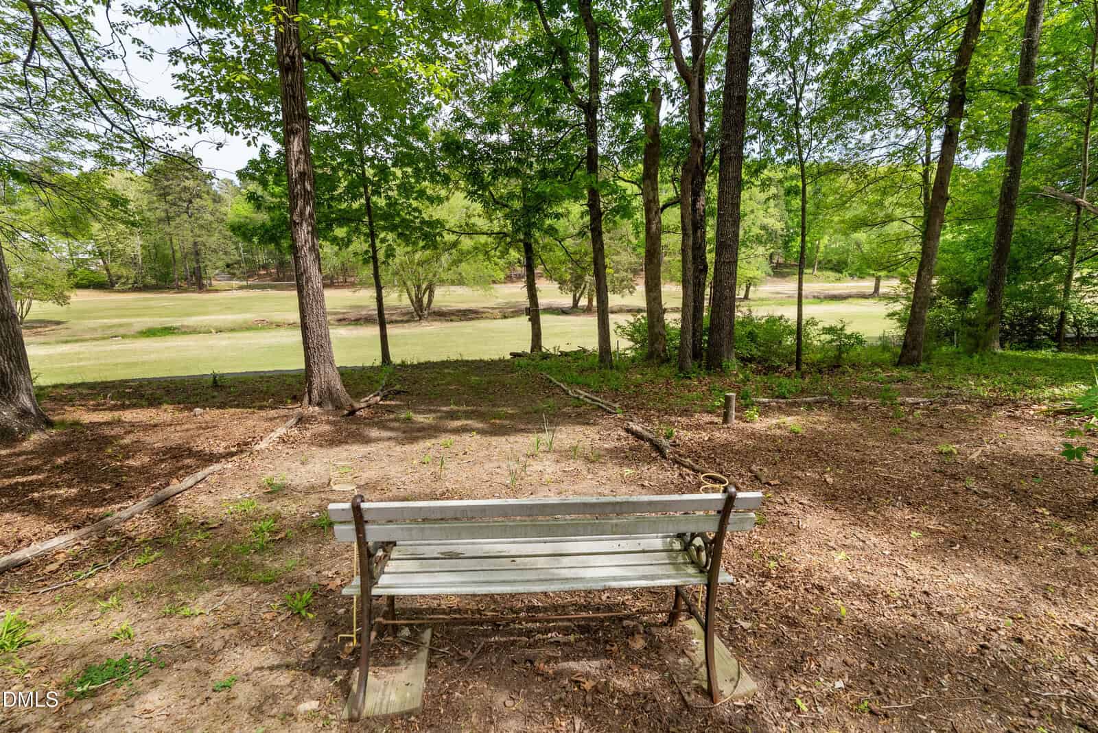 A wooden bench sits in a shaded, wooded area overlooking a grassy field dotted with trees under a clear sky.