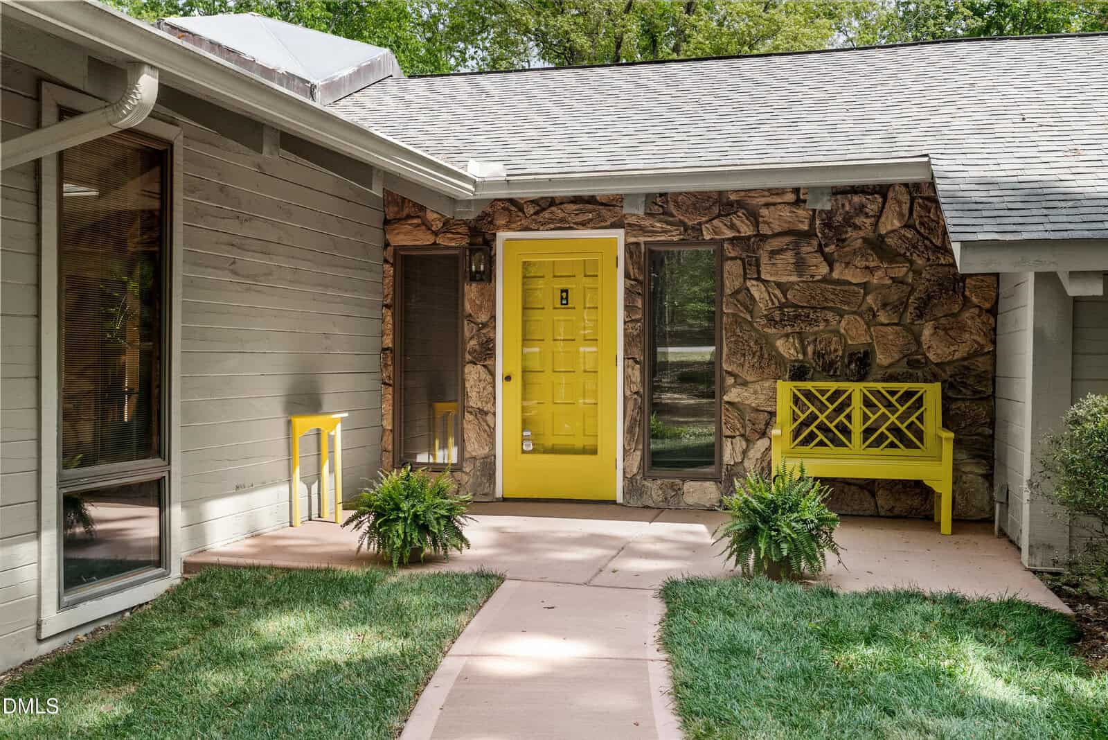 A home entryway with a bold yellow door and matching bench, stone and wood exterior walls, large windows, potted ferns, curved path, and neatly trimmed green lawn.