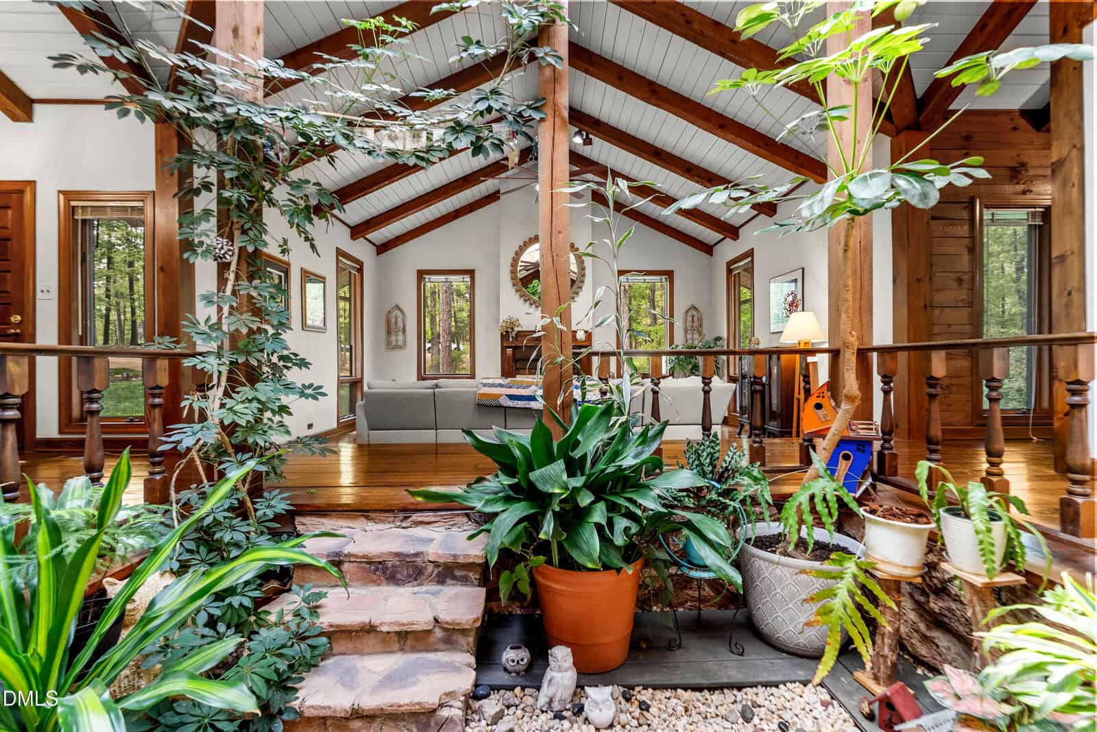 Bright living space with exposed beams, large plants, stone steps, wood floors, and many windows letting in natural light. Cozy seating and decor create a warm, inviting atmosphere connecting indoors with nature.