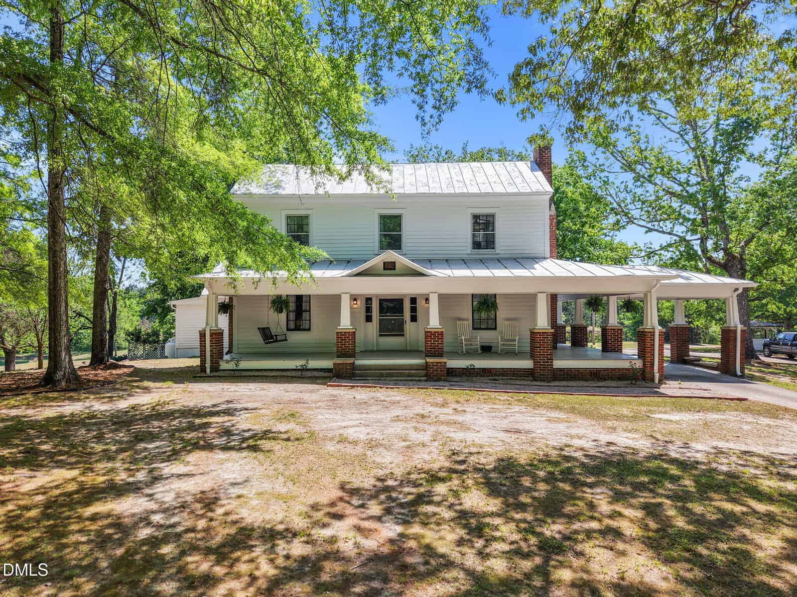 A large two-story white farmhouse with a metal roof and a spacious wraparound porch, surrounded by tall green trees and a dirt yard under a bright blue sky.