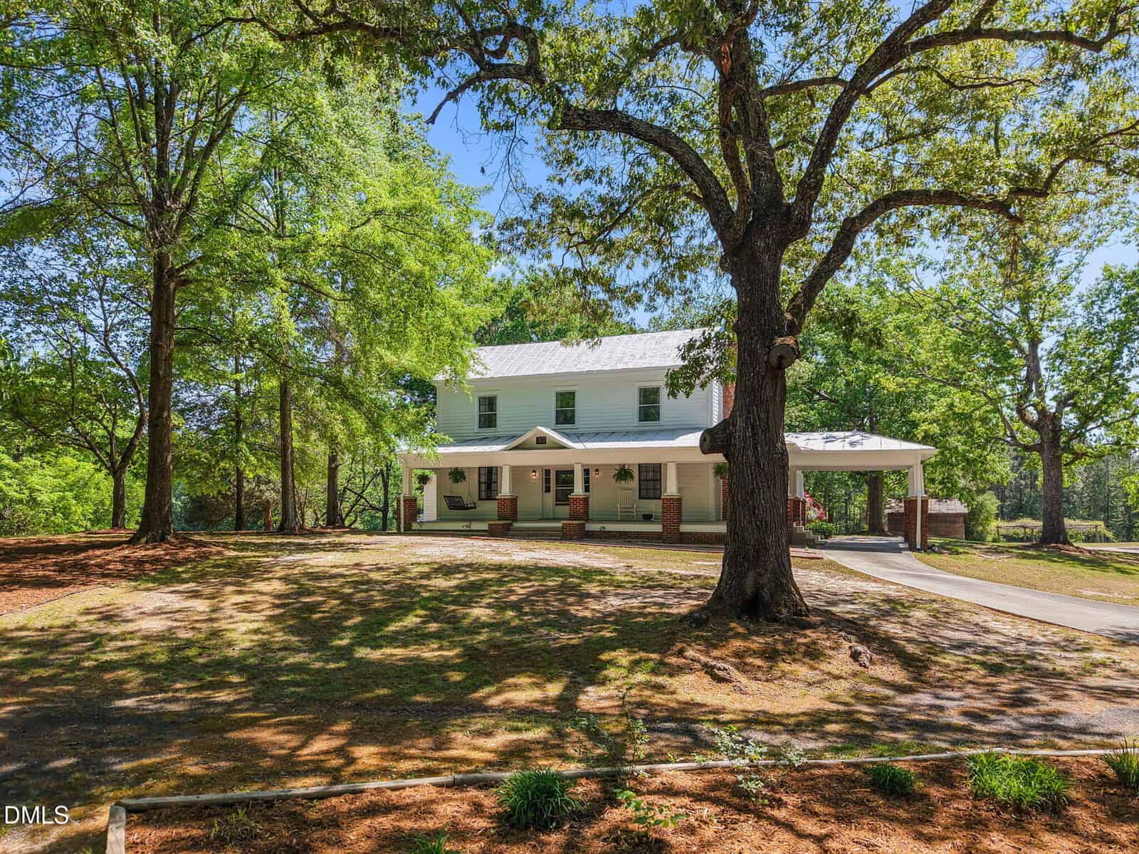 A two-story white house with a large front porch sits among tall trees and a shaded, spacious yard on a sunny day. A driveway leads to a carport on the right side of the house.